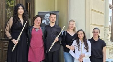 Six people pose on stone steps outside a building, each holding a flute, with a portrait poster in the background.