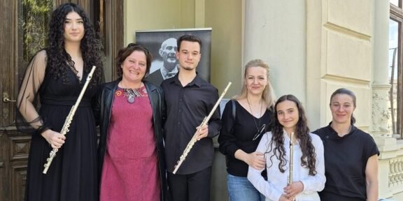Six people pose on stone steps outside a building, each holding a flute, with a portrait poster in the background.