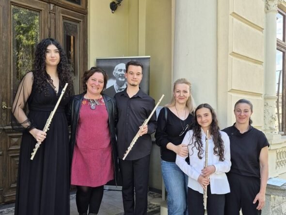 Six people pose on stone steps outside a building, each holding a flute, with a portrait poster in the background.