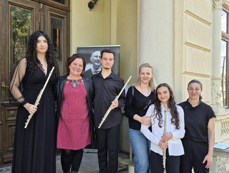 Six people pose on stone steps outside a building, each holding a flute, with a portrait poster in the background.