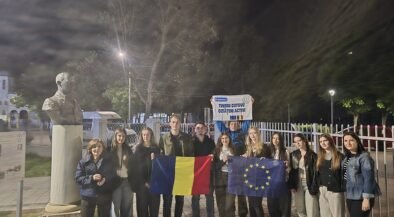 Group of young people posing at night, holding Romanian and EU flags in front of a statue bust and fence outside a building.