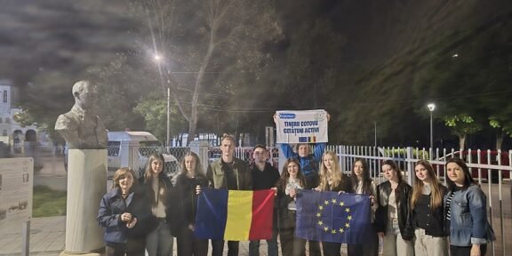 Group of young people posing at night, holding Romanian and EU flags in front of a statue bust and fence outside a building.