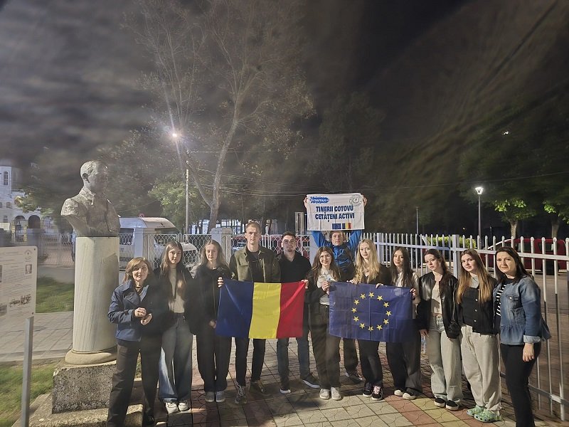 Group of young people posing at night, holding Romanian and EU flags in front of a statue bust and fence outside a building.
