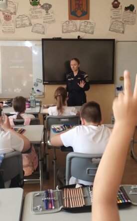 Uniformed officer giving a talk to elementary students in a classroom; a student in the foreground raises a hand to answer.