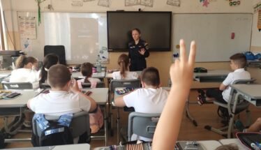 Uniformed officer giving a talk to elementary students in a classroom; a student in the foreground raises a hand to answer.