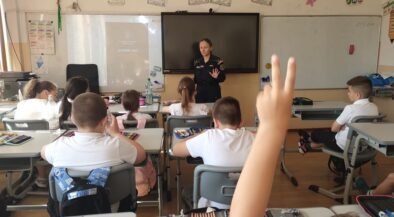 Uniformed officer giving a talk to elementary students in a classroom; a student in the foreground raises a hand to answer.