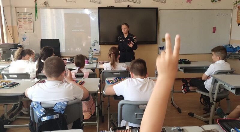 Uniformed officer giving a talk to elementary students in a classroom; a student in the foreground raises a hand to answer.