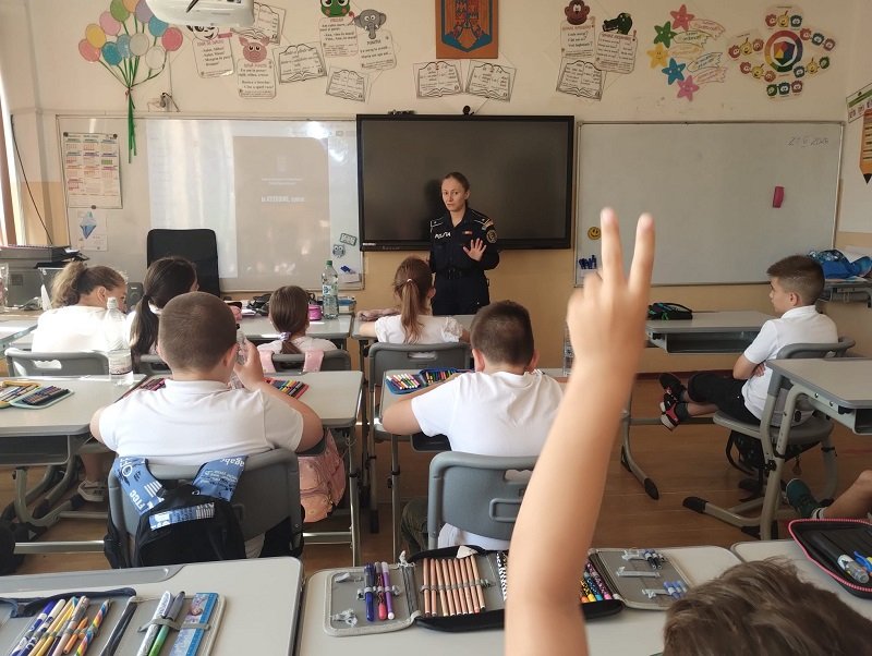 Uniformed officer giving a talk to elementary students in a classroom; a student in the foreground raises a hand to answer.
