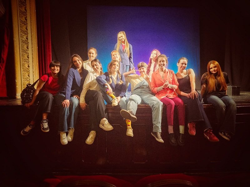 A group of about 12 young people posing together on a theater stage, some sitting on the edge and one standing center, smiling at the camera.
