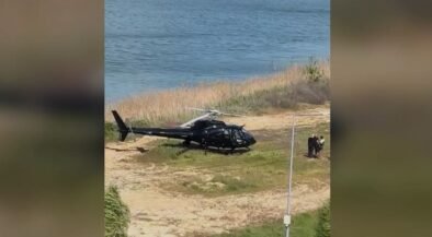 Black helicopter parked on a sandy area by the water, with a few people standing nearby.
