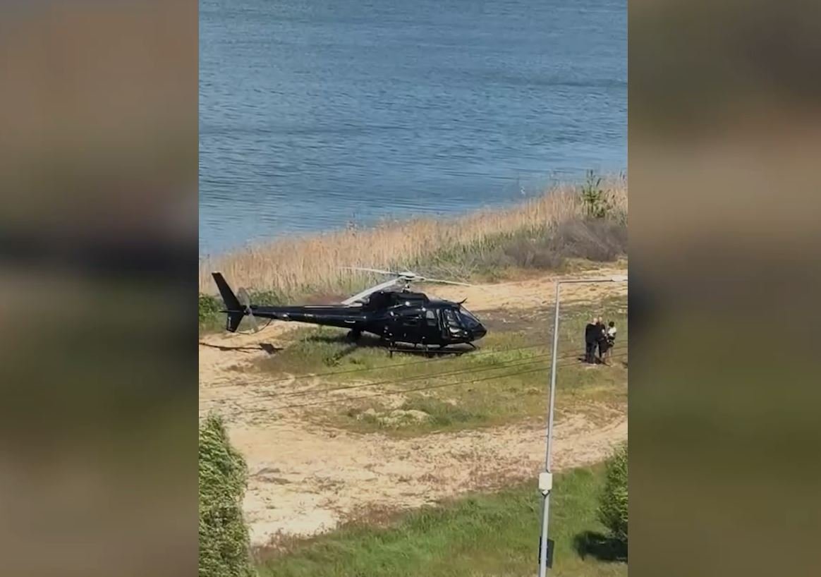 Black helicopter parked on a sandy area by the water, with a few people standing nearby.