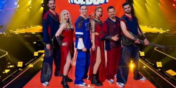 A group of performers posing on a brightly lit stage with musical instruments and red carpet, wearing colorful costumes.