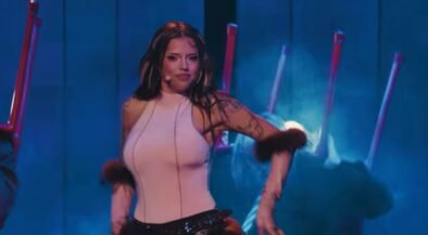 Female performer on stage wearing a pale pink sleeveless top with fur cuffs, headset microphone, blue lighting, and backup dancers in the background.