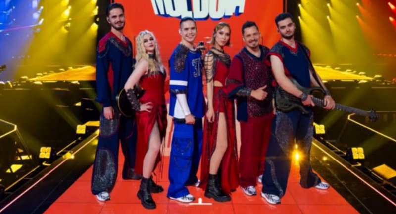 A group of performers posing on a brightly lit stage with musical instruments and red carpet, wearing colorful costumes.