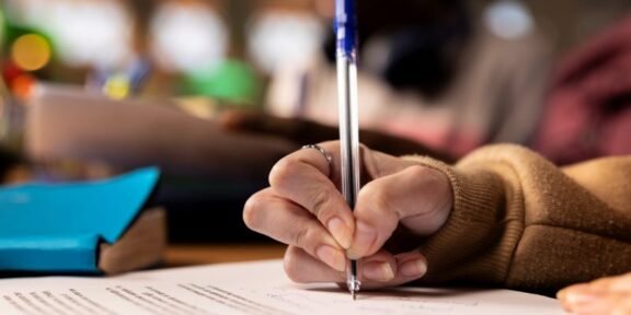Close-up of a hand in a brown sleeve writing with a blue pen on a printed document at a desk.