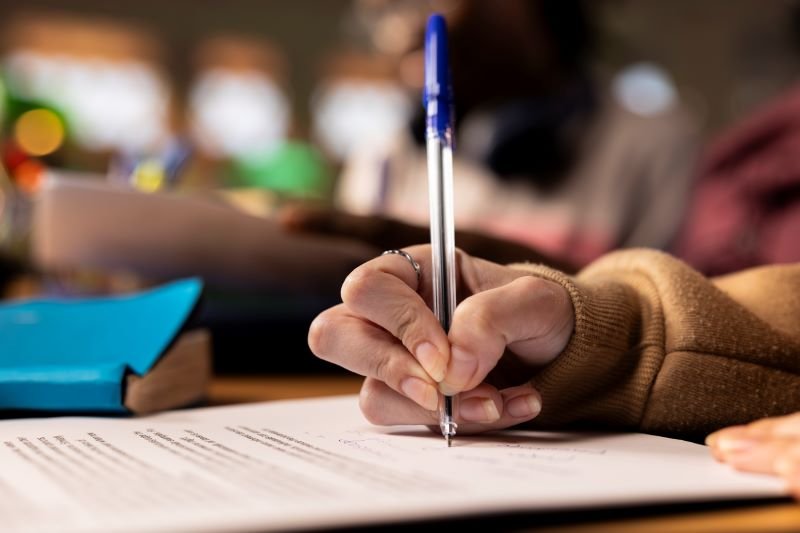 Close-up of a hand in a brown sleeve writing with a blue pen on a printed document at a desk.