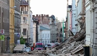 Urban street after a structural collapse, with a large pile of rubble blocking the road and damaged buildings on both sides, several vehicles in the background.