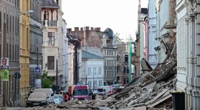 Urban street after a structural collapse, with a large pile of rubble blocking the road and damaged buildings on both sides, several vehicles in the background.