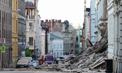 Urban street after a structural collapse, with a large pile of rubble blocking the road and damaged buildings on both sides, several vehicles in the background.