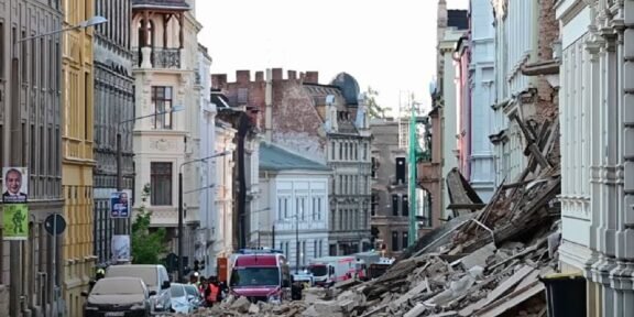 Urban street after a structural collapse, with a large pile of rubble blocking the road and damaged buildings on both sides, several vehicles in the background.