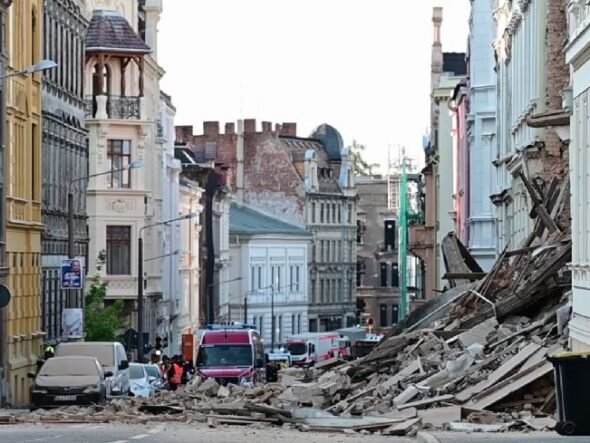 Urban street after a structural collapse, with a large pile of rubble blocking the road and damaged buildings on both sides, several vehicles in the background.