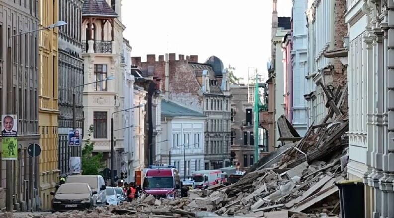 Urban street after a structural collapse, with a large pile of rubble blocking the road and damaged buildings on both sides, several vehicles in the background.