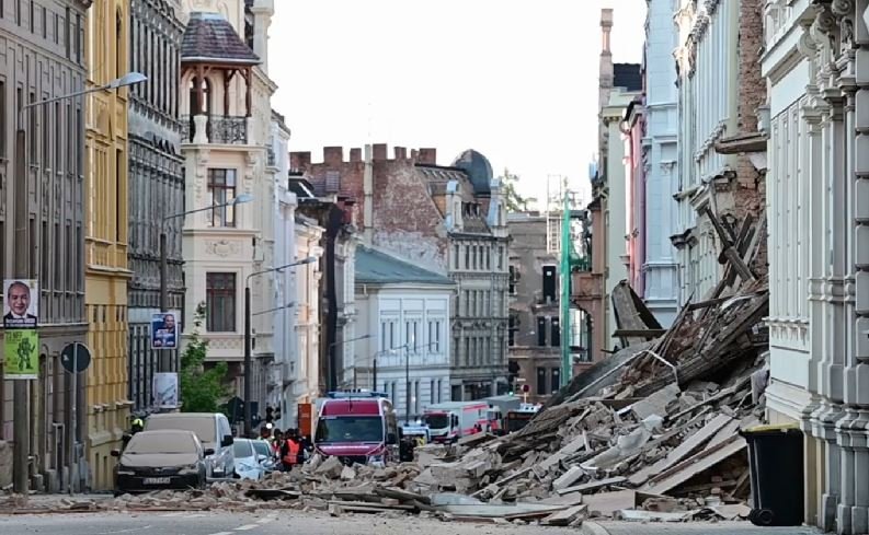 Urban street after a structural collapse, with a large pile of rubble blocking the road and damaged buildings on both sides, several vehicles in the background.
