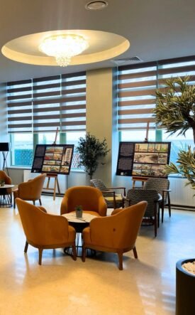 Lounge-like room with orange chairs around tables, display boards showing information, and potted plants by large windows; a blue branded panel with neon logo on the right.