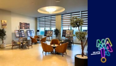 Lounge-like room with orange chairs around tables, display boards showing information, and potted plants by large windows; a blue branded panel with neon logo on the right.