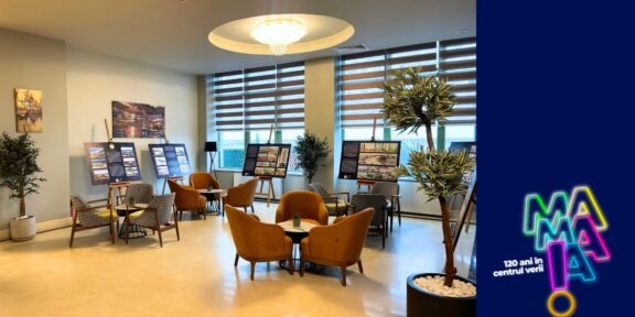 Lounge-like room with orange chairs around tables, display boards showing information, and potted plants by large windows; a blue branded panel with neon logo on the right.
