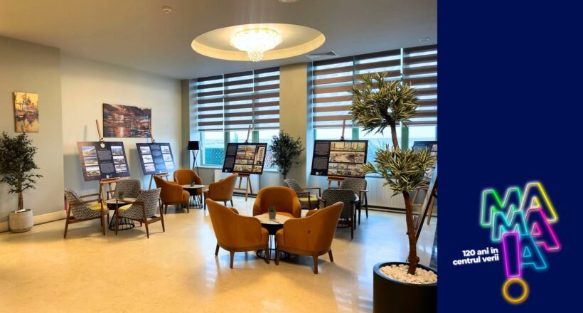 Lounge-like room with orange chairs around tables, display boards showing information, and potted plants by large windows; a blue branded panel with neon logo on the right.