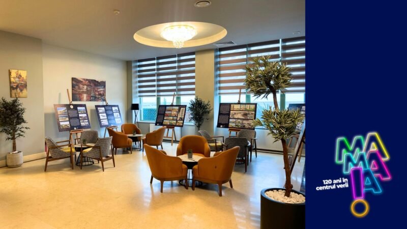 Lounge-like room with orange chairs around tables, display boards showing information, and potted plants by large windows; a blue branded panel with neon logo on the right.