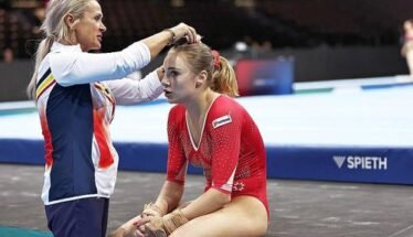 Coach ties a gymnast's hair before a routine, both seated on a competition mat in a gym.