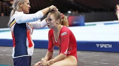 Coach ties a gymnast's hair before a routine, both seated on a competition mat in a gym.