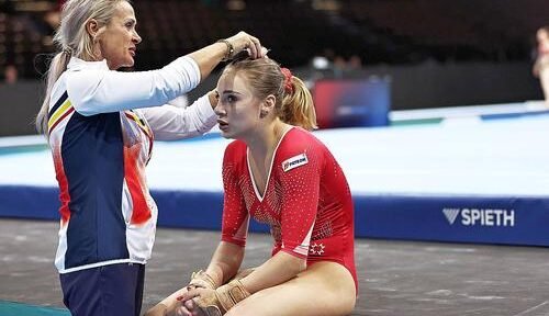 Coach ties a gymnast's hair before a routine, both seated on a competition mat in a gym.