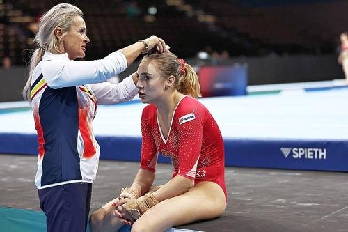 Coach ties a gymnast's hair before a routine, both seated on a competition mat in a gym.
