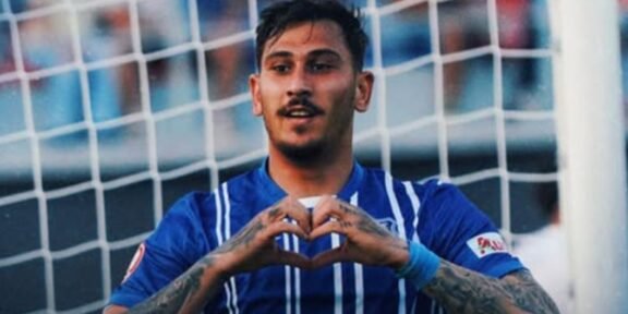 Soccer player in a blue jersey forming a heart shape with his hands in front of a goal net.