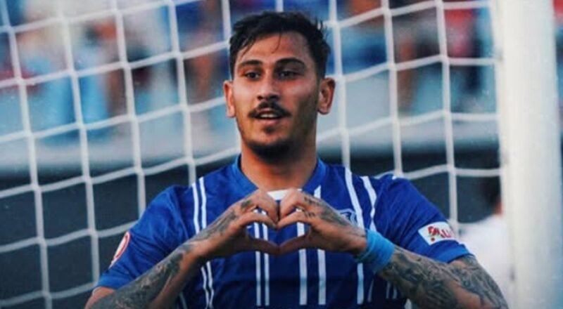 Soccer player in a blue jersey forming a heart shape with his hands in front of a goal net.