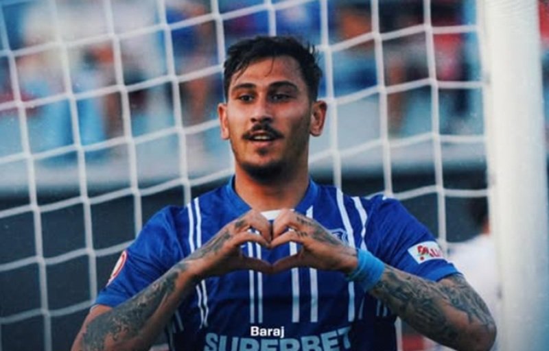 Soccer player in a blue jersey forming a heart shape with his hands in front of a goal net.