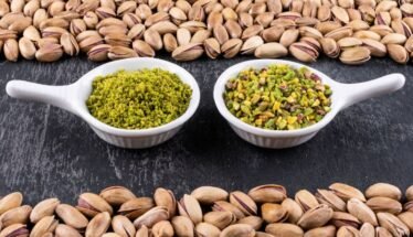 Two white spoons sit on a dark surface, each filled with pistachio-based spreads: green pesto on the left and chopped pistachios on the right, framed by pistachio shells around the edge of the frame.