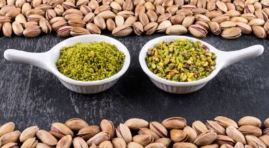 Two white spoons sit on a dark surface, each filled with pistachio-based spreads: green pesto on the left and chopped pistachios on the right, framed by pistachio shells around the edge of the frame.