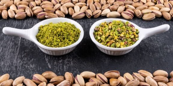Two white spoons sit on a dark surface, each filled with pistachio-based spreads: green pesto on the left and chopped pistachios on the right, framed by pistachio shells around the edge of the frame.