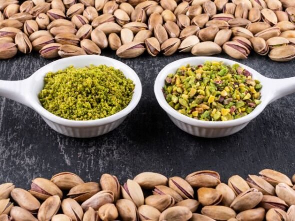 Two white spoons sit on a dark surface, each filled with pistachio-based spreads: green pesto on the left and chopped pistachios on the right, framed by pistachio shells around the edge of the frame.