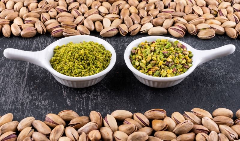 Two white spoons sit on a dark surface, each filled with pistachio-based spreads: green pesto on the left and chopped pistachios on the right, framed by pistachio shells around the edge of the frame.
