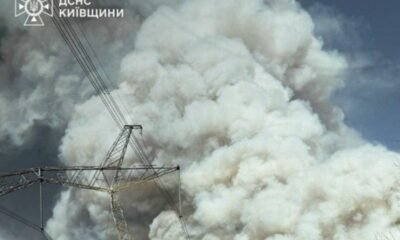Massive white smoke plume behind a high-voltage transmission tower and lines; emergency services logo in the top-left corner.