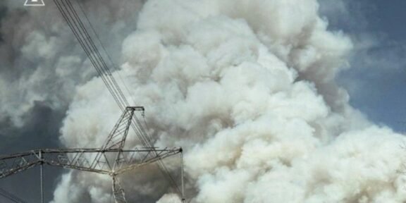 Massive white smoke plume behind a high-voltage transmission tower and lines; emergency services logo in the top-left corner.