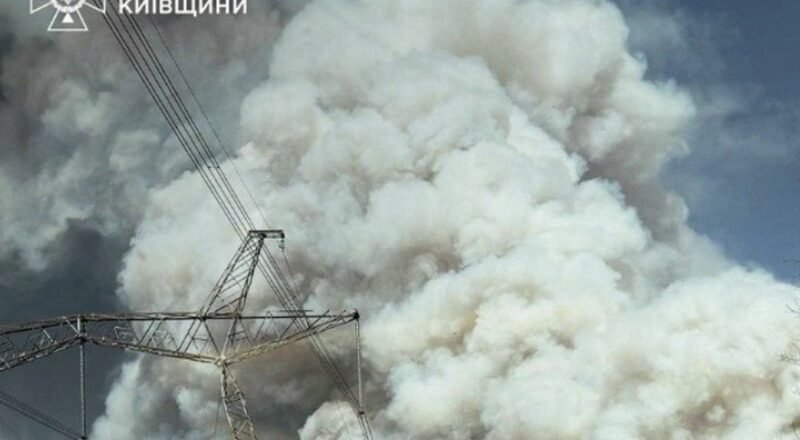 Massive white smoke plume behind a high-voltage transmission tower and lines; emergency services logo in the top-left corner.
