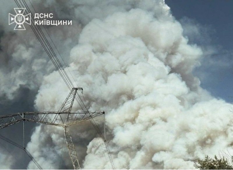 Massive white smoke plume behind a high-voltage transmission tower and lines; emergency services logo in the top-left corner.