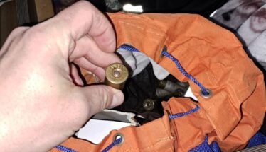 Close-up of a hand holding a brass shotgun shell over an orange bag with blue cords; inside are more shells, Romanian police logo visible