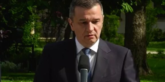 Man in a dark suit speaks into a microphone outdoors, with trees in the background.